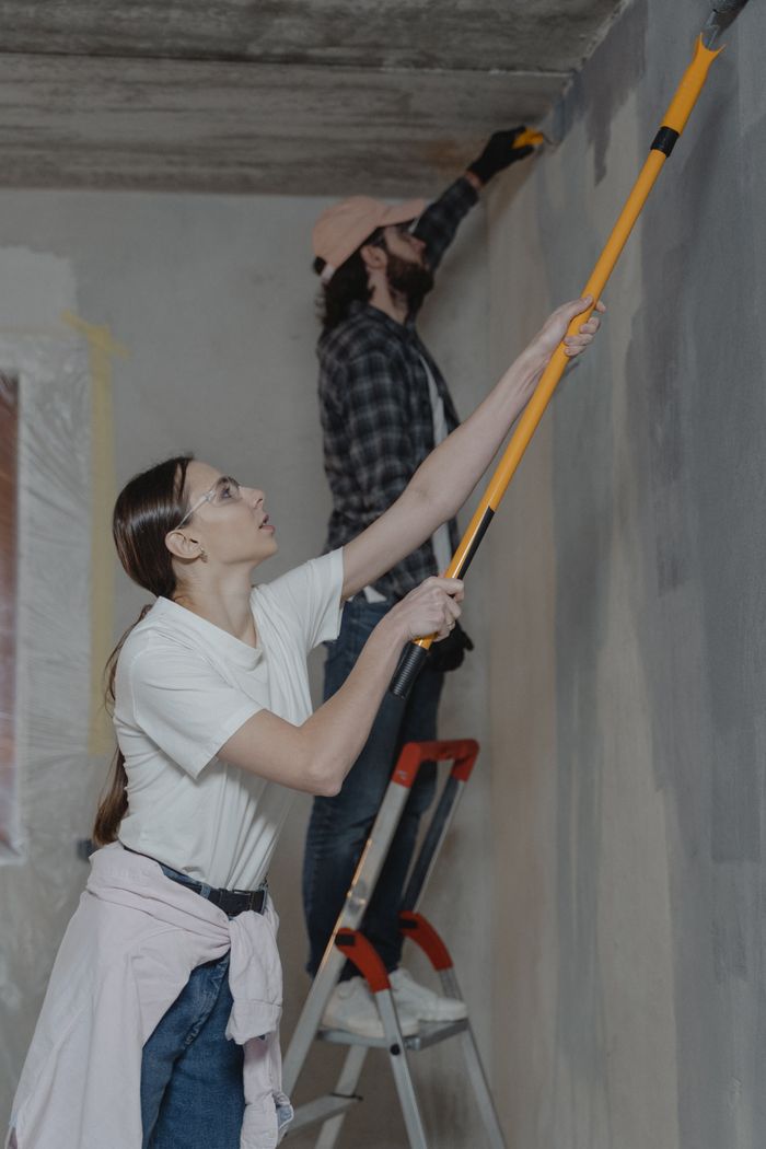 A couple painting a room wall during home renovation. Both are actively working with tools.